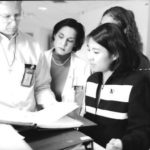 1 man and 3 women, all doctors, stand looking at a medical chart, black and white image.